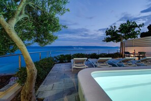 Area piscina con sedie a sdraio e un tavolo, con vista su una distesa d'acqua e una costa lontana.