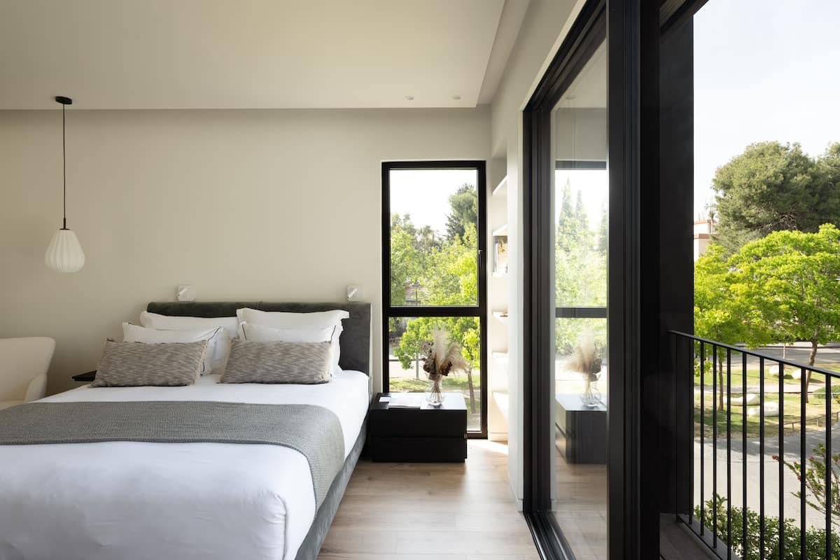 A modern bedroom with a large bed, a bedside table with a vase, and a view of greenery outside.