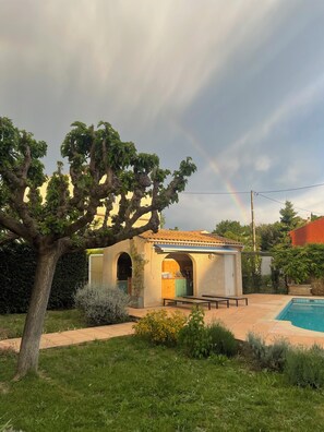 Een huis met een zwembad, een boom en een regenboog aan de lucht.