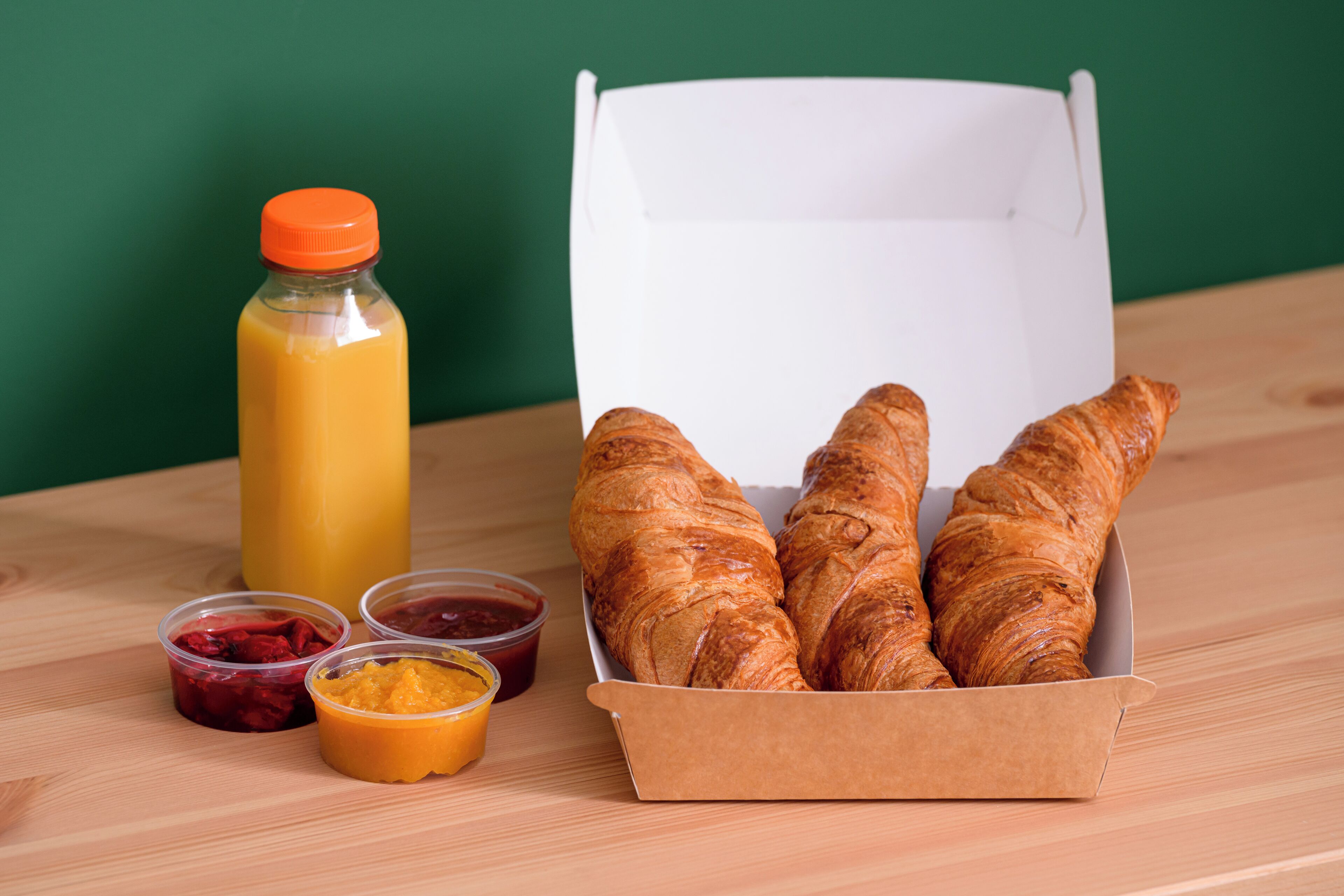 A box of croissants, a bottle of orange juice, and two containers of jam on a wooden table.