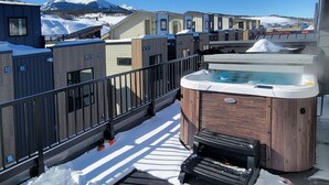 A hot tub on a balcony with snow-covered ground, surrounded by modern buildings with wooden facades.