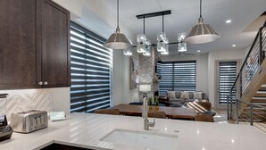 A modern kitchen with a white countertop, dark cabinets, and a built-in toaster. There is a dining area with a wooden table and chairs, and a staircase leading to an upper level.