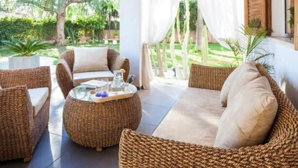A sunlit outdoor seating area with wicker furniture and a glass-top table, overlooking a lush garden.