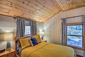Quarto acolhedor com teto de madeira, cama com colcha amarela, duas luminárias de cabeceira e uma janela com cortinas.