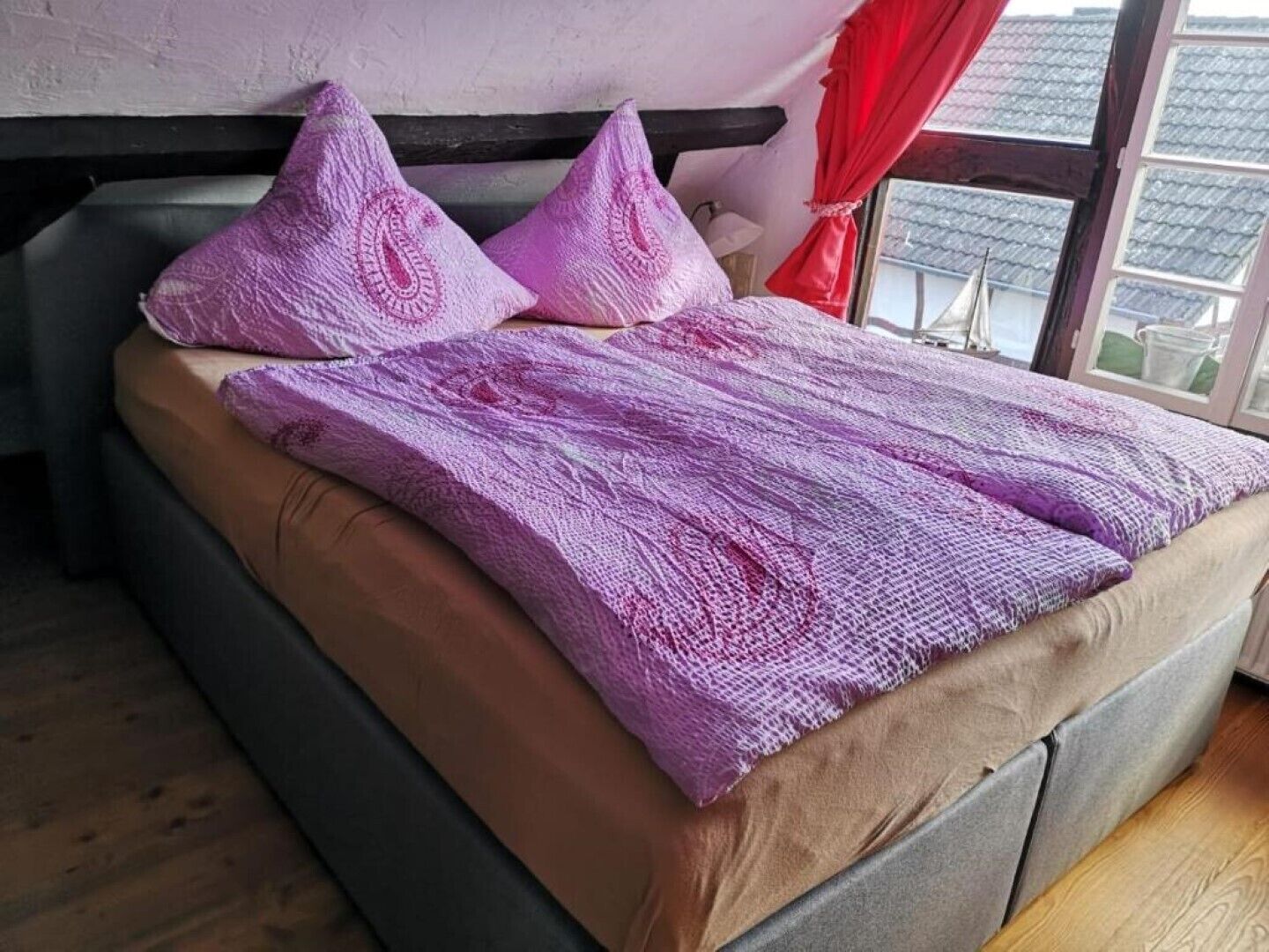 Un lit avec une couverture et des oreillers violets, une fenêtre donnant sur des immeubles et un rideau rouge.