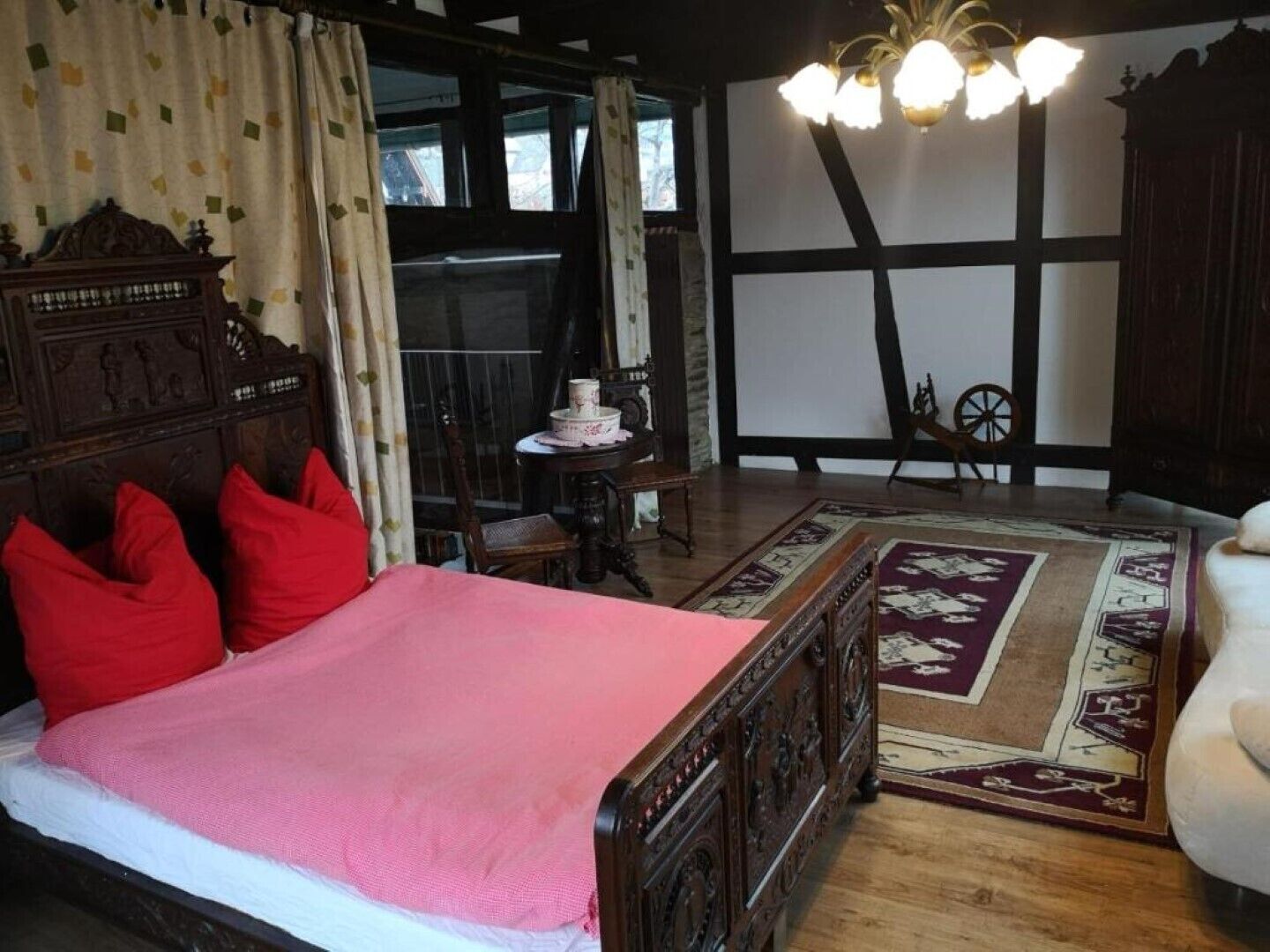 Ein traditionell eingerichteter Raum mit einem großen Bett, einem Holzkopfende, einer Chandelierleuchtre und Blick durch ein mit Vorhängen drapiertes Fenster nach draußen.
