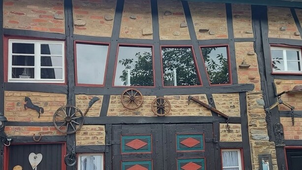 Il s’agit d’un bâtiment traditionnel à colombages, doté d’une porte et de fenêtres noires, d’un banc en bois et d’une table sur laquelle est posée une plante en pot.