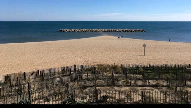 Ein sandiger Strand mit einem hölzernen Zaun im Vordergrund und einem Felsstollen im Hintergrund.