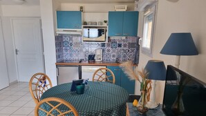 Eine kompakte Küche mit blauen Schränken, einer Mikrowelle und einer Kaffeemaschine. Ein Esstisch mit Stühlen und ein Fernseher auf einem Lowboard.