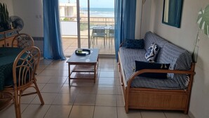 Ein Wohnzimmer mit einer Couch, einem Tisch und einem Sessel. Durch eine Glastür ist der Blick auf den Ozean.