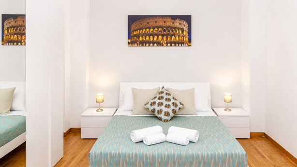 A hotel room with a bed, two bedside tables, a nightstand, a pillow, a blanket, and a wall-mounted picture of the Colosseum.