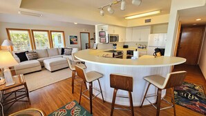 A modern kitchen with a central island, bar stools, and a dining area with a sofa and lamp. - Beachfront - Community Pool/Pickleball Access! (St. George Island)