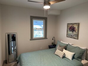 A bedroom with a bed, a ceiling fan, a window, a nightstand, a lamp, and a framed picture on the wall.