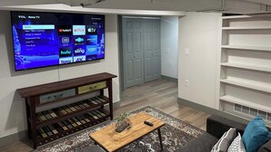 Ein Wohnzimmer mit Flachbildfernseher, der Streaming-Dienste anzeigt, einem Holztisch, einer Couch und einem Bücherregal.