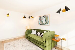 Un canapé vert agrémenté de coussins à motifs, une petite table avec une lampe et un tableau accroché au mur.