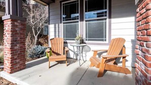 Un porche abrité avec deux fauteuils Adirondack et une petite table, une colonne en briques et une fenêtre avec des stores.