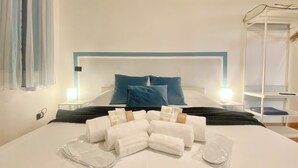 Un llit ben fet amb almoades blaves i blanques, una taula de nit amb una làmpada, i un conjunt de tovalloles i productes de tocador damunt del llit.