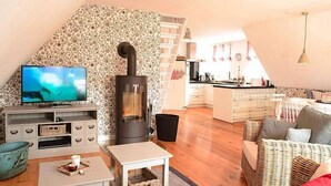 A cozy living room with a fireplace, a television, and a kitchen area in the background. - Ferienwohnung in Wyk auf Foehr (Wyk auf Föhr)