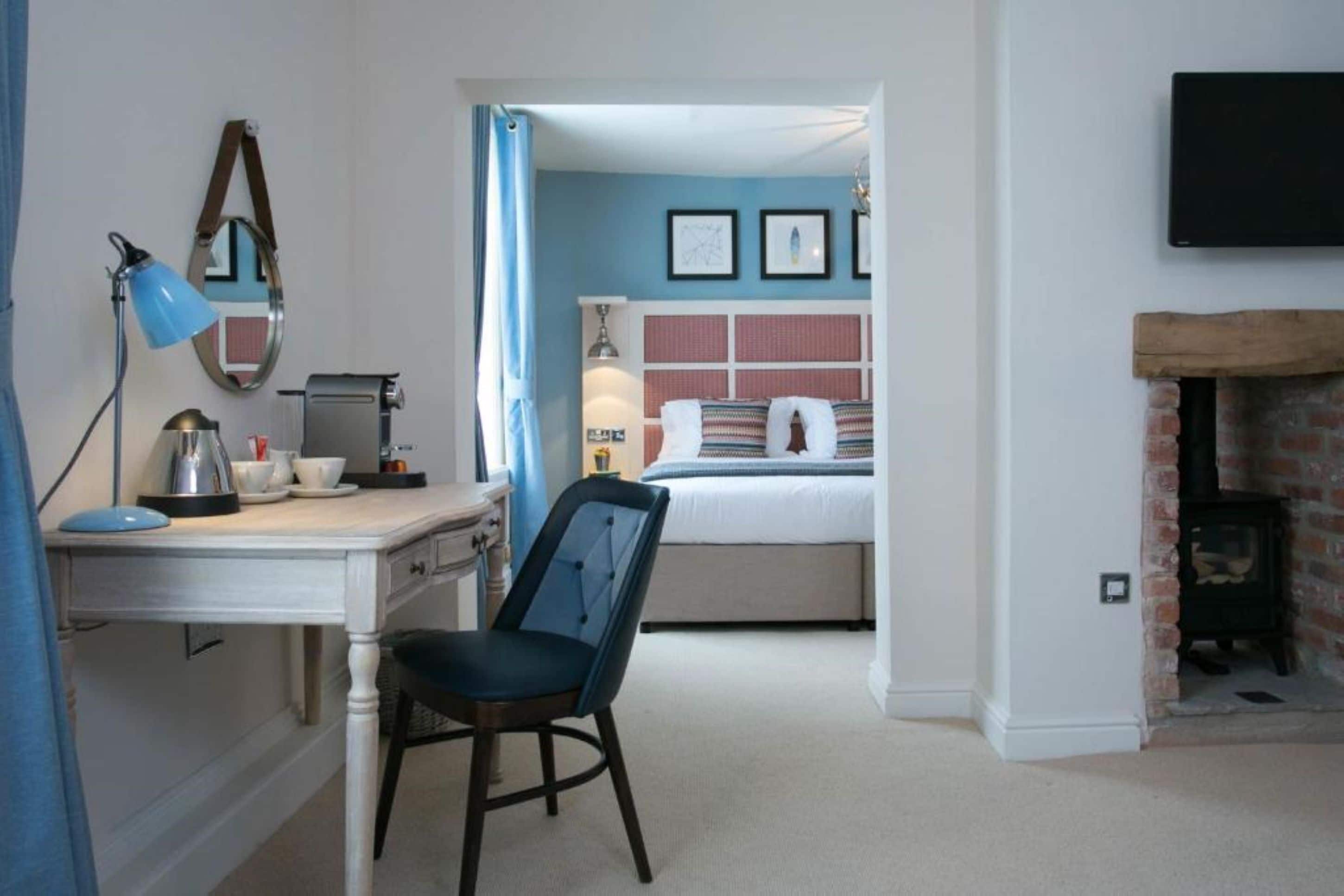 A bedroom with a bed, a desk with a lamp, a chair, and a fireplace.