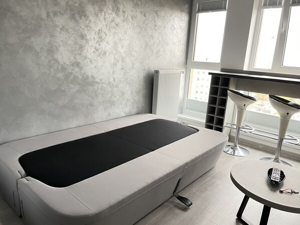 A modern room with a minimalist design, featuring a single bed, a small round table, and a window with blinds.