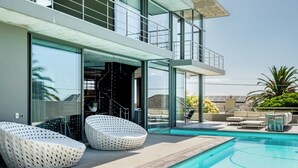 A modern outdoor pool area with two lounge chairs, a glass-enclosed entrance, and a view of the surrounding landscape.