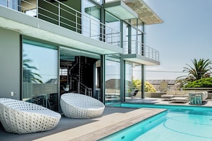 A modern outdoor pool area with two lounge chairs, a glass-enclosed entrance, and a view of the surrounding landscape.