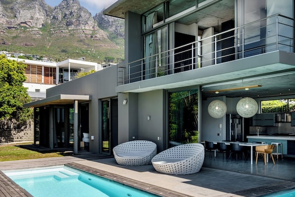 A modern two-story house with a swimming pool, outdoor seating, and a view of mountains.