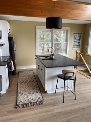 A modern kitchen with a central island, a hanging light fixture, and a view of trees through a large window.