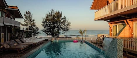 A pool with a view of the ocean, wooden deck chairs, and a wooden building with a balcony.