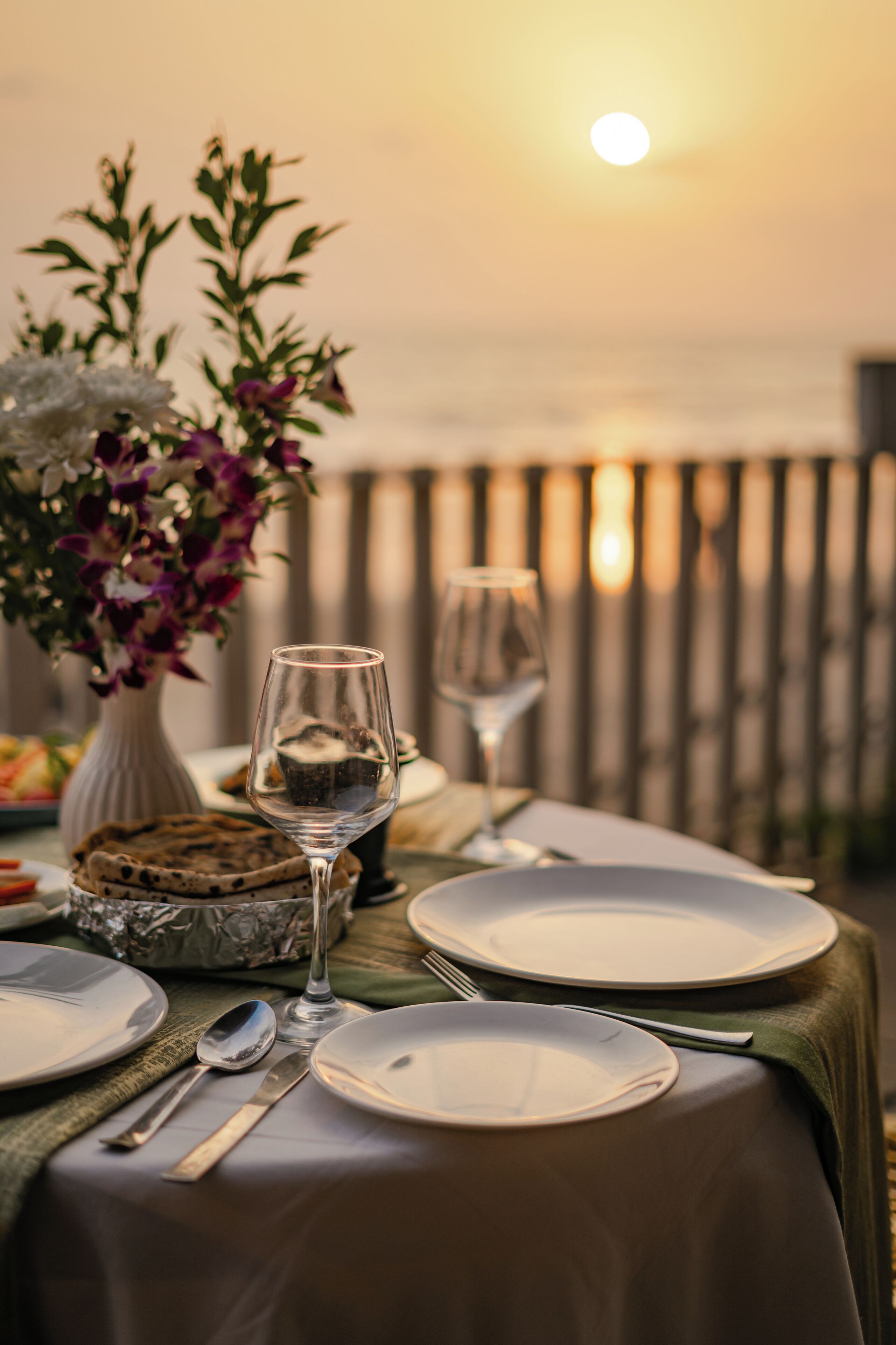 A table set for two with plates, cutlery, and a glass, positioned near a railing with a view of the sunset.