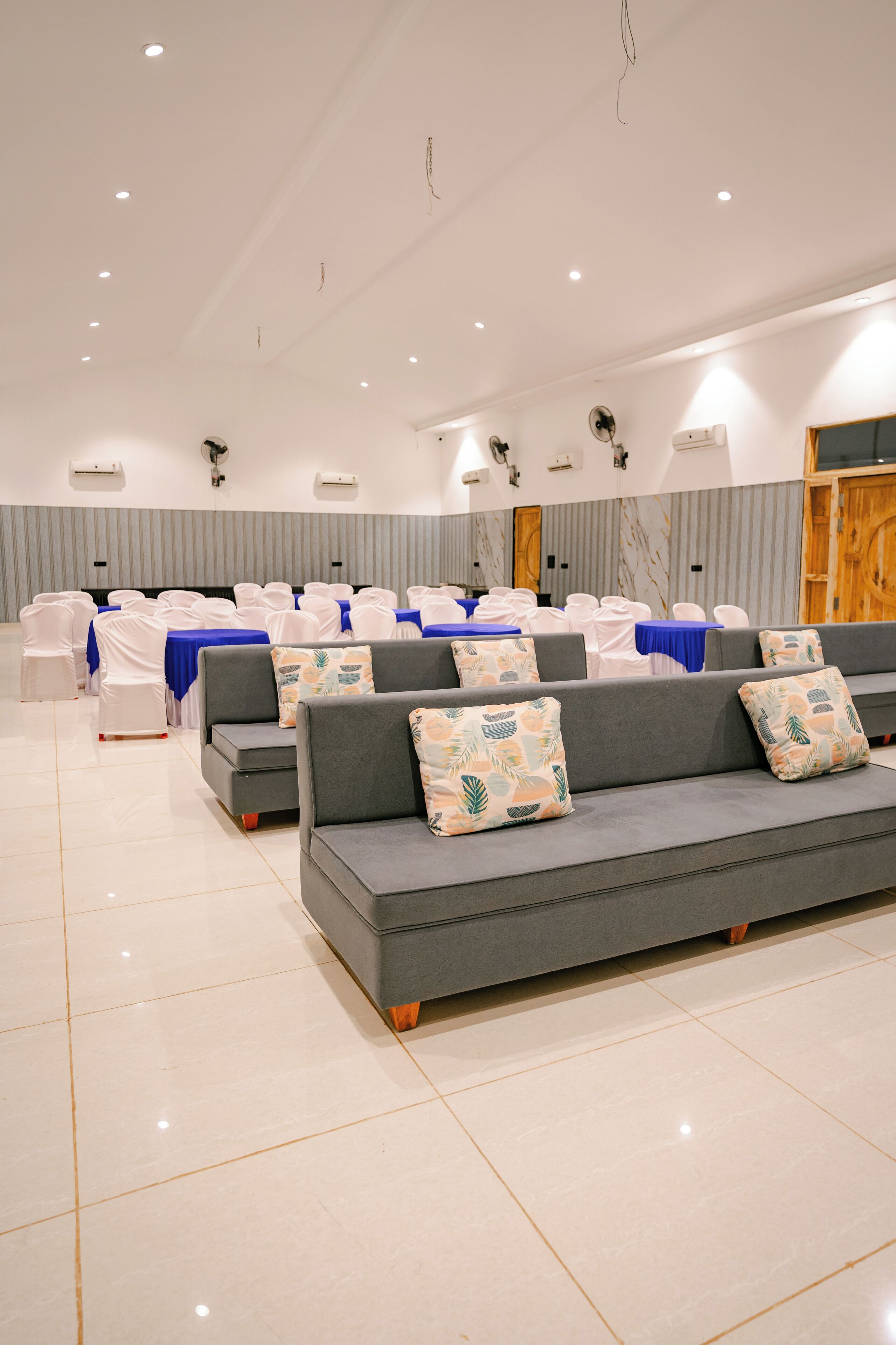 Une salle de conférence spacieuse, avec des rangées de chaises blanches, un canapé gris orné de coussins à motifs et une table recouverte d’une nappe bleue.