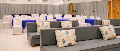 Une salle de conférence spacieuse, avec des rangées de chaises blanches, un canapé gris orné de coussins à motifs et une table recouverte d’une nappe bleue.