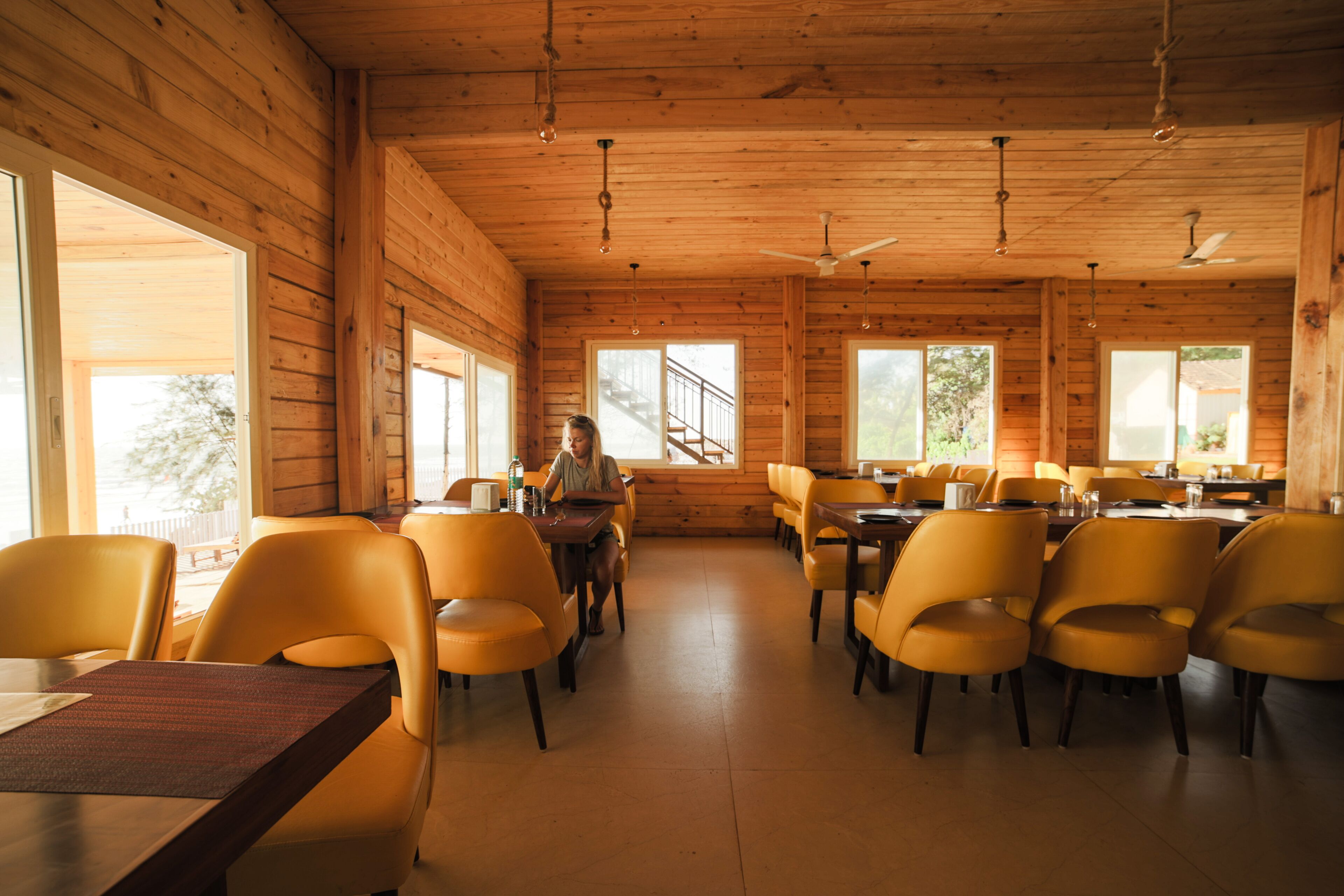 A wooden interior with tables and yellow chairs, large windows, and a staircase.