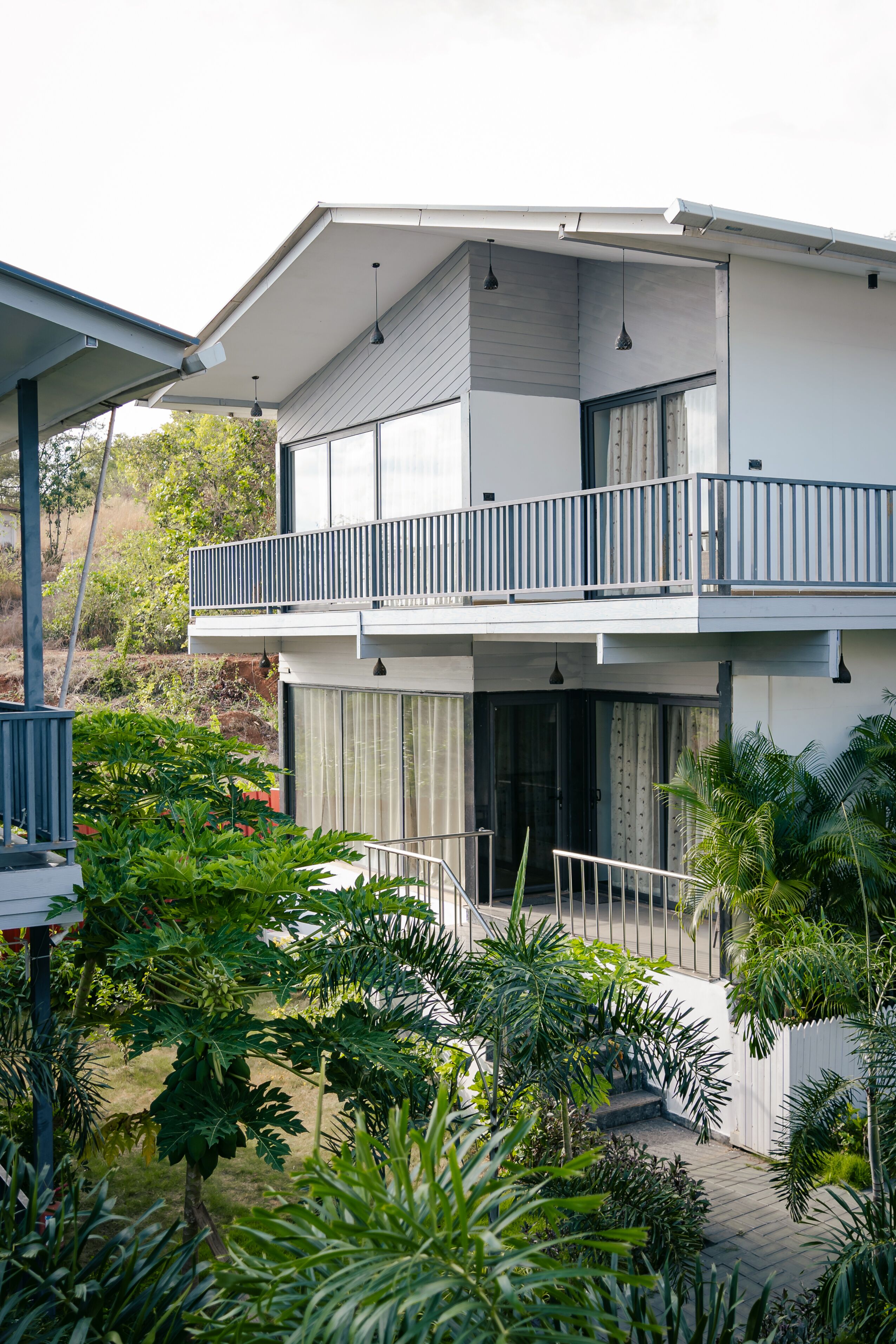 A modern two-story house with a balcony, surrounded by lush greenery.