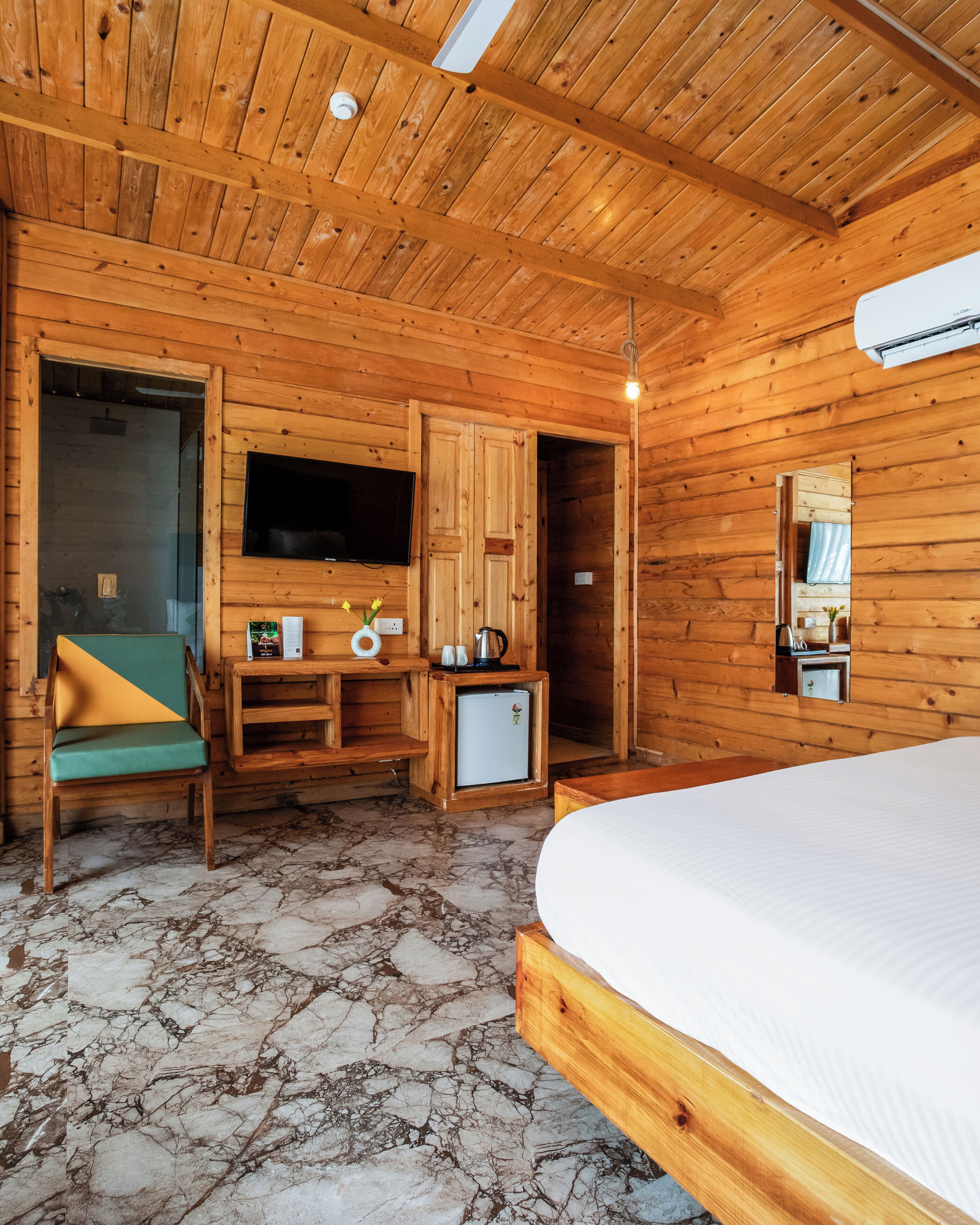 A wooden cabin room with a bed, a TV, a small fridge, a wooden chair, and a wooden desk with a kettle.