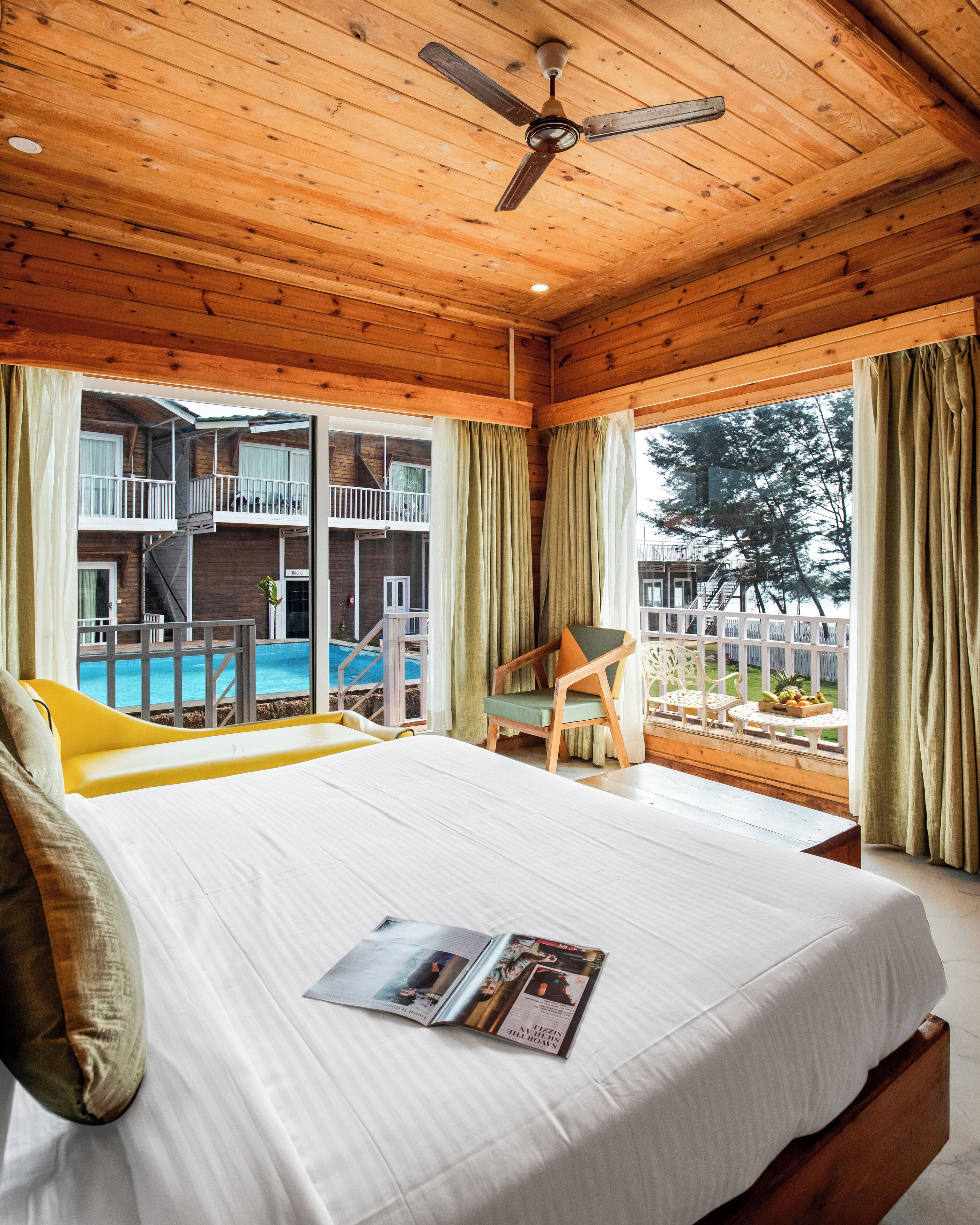 A wooden cabin with a bed, a ceiling fan, a balcony with a view of a pool and trees.