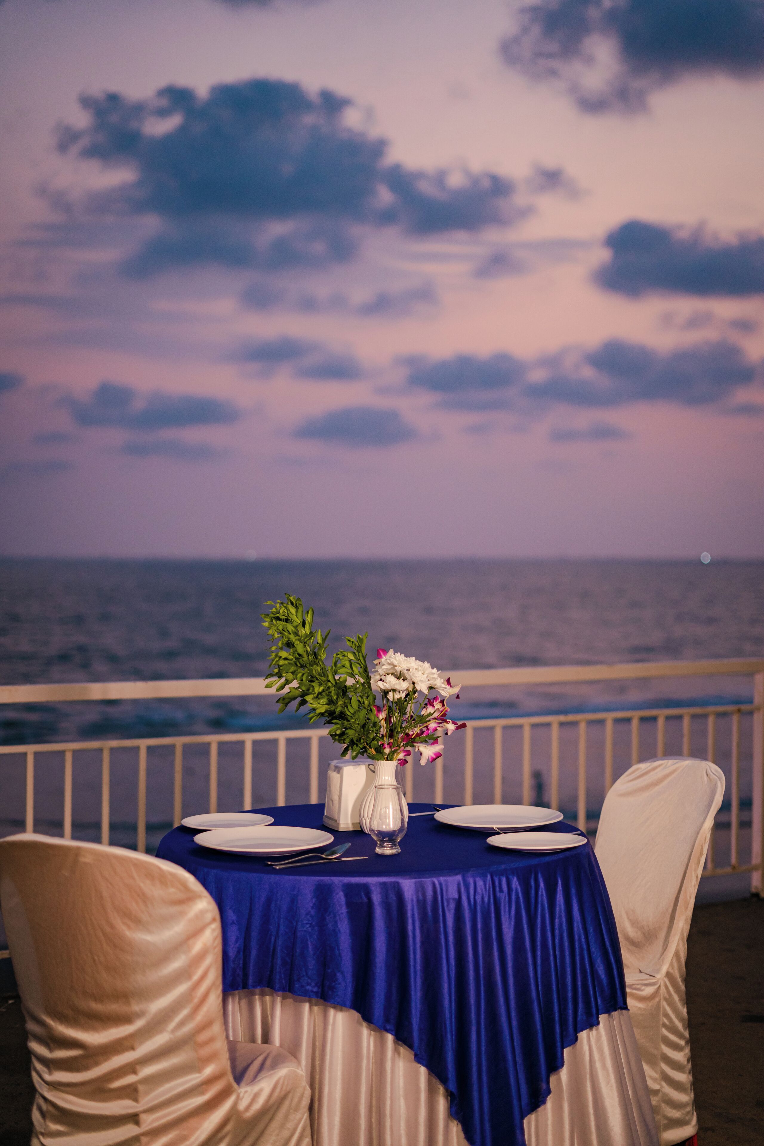 Mesa para dos con mantel azul, platos blancos y un jarrón de flores, con vista al océano.