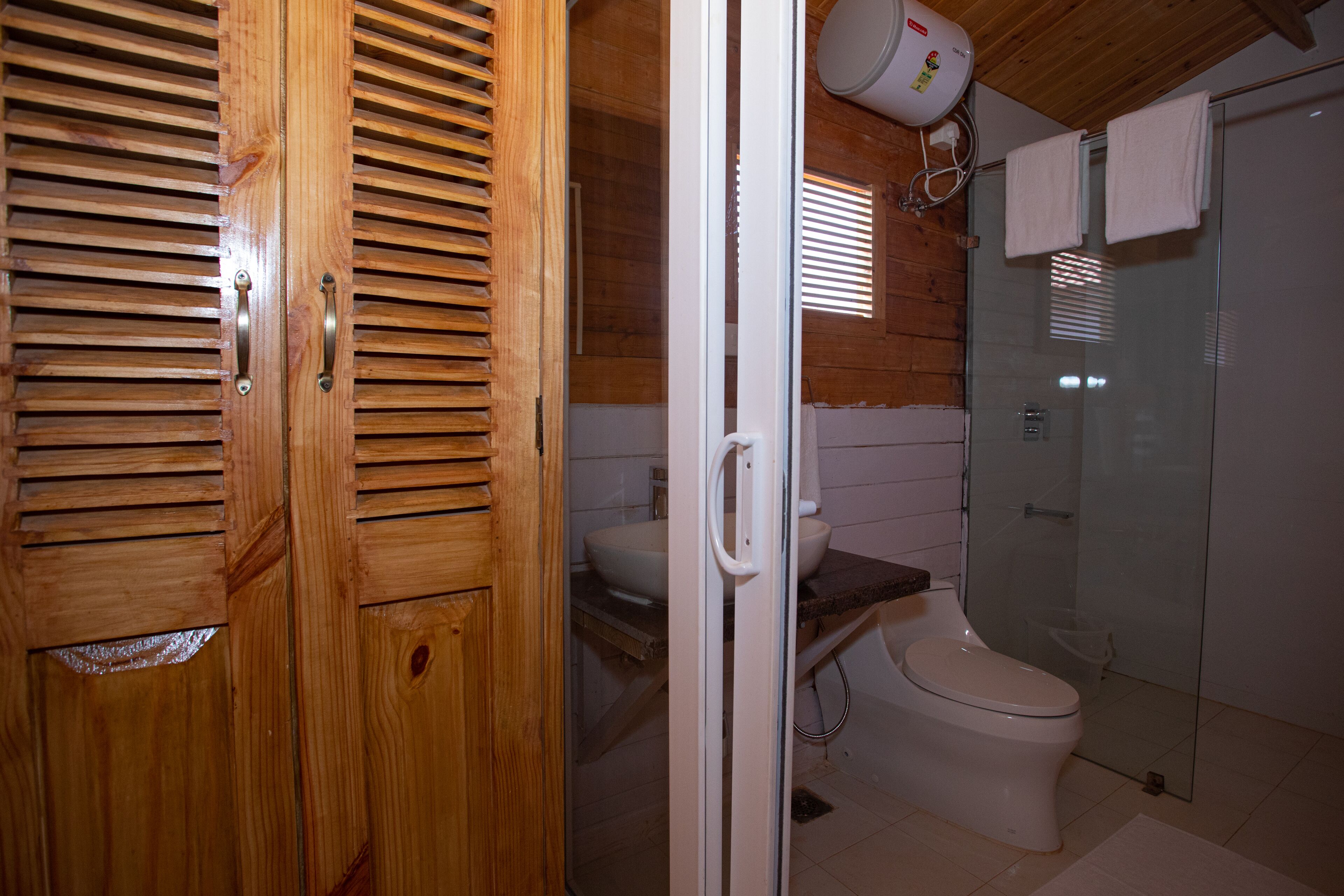 Une porte en bois à panneaux ajourés, des toilettes blanches, une cabine de douche en verre et une serviette blanche accrochée au mur.