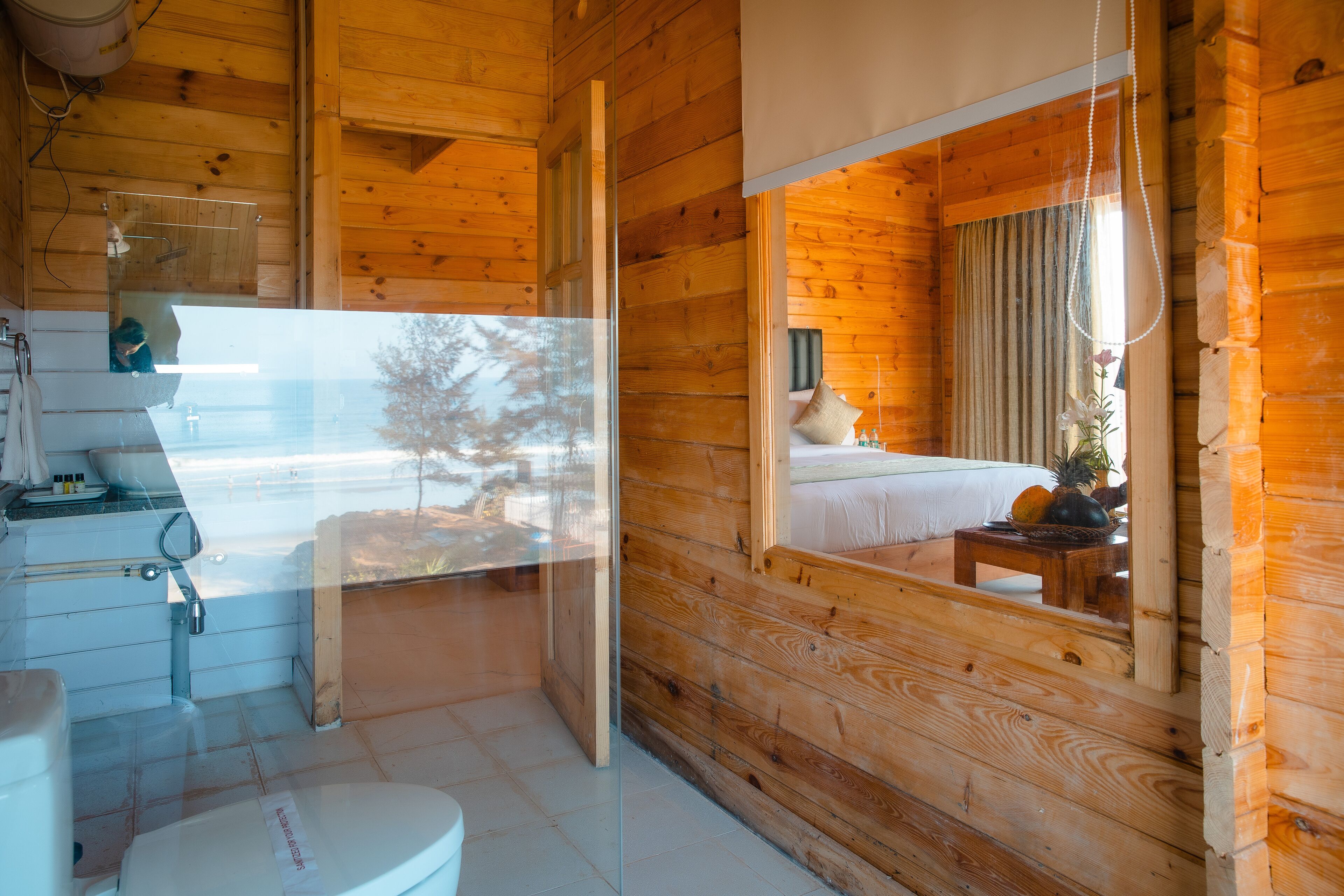 A wooden cabin interior with a bathroom, a bed, and a window with a view of trees.