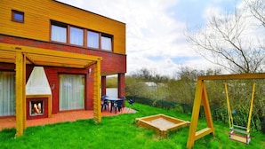 Une maison moderne à deux étages, avec une façade en briques et une terrasse en bois. On y trouve un espace patio avec une table et des chaises, ainsi qu’une balançoire.