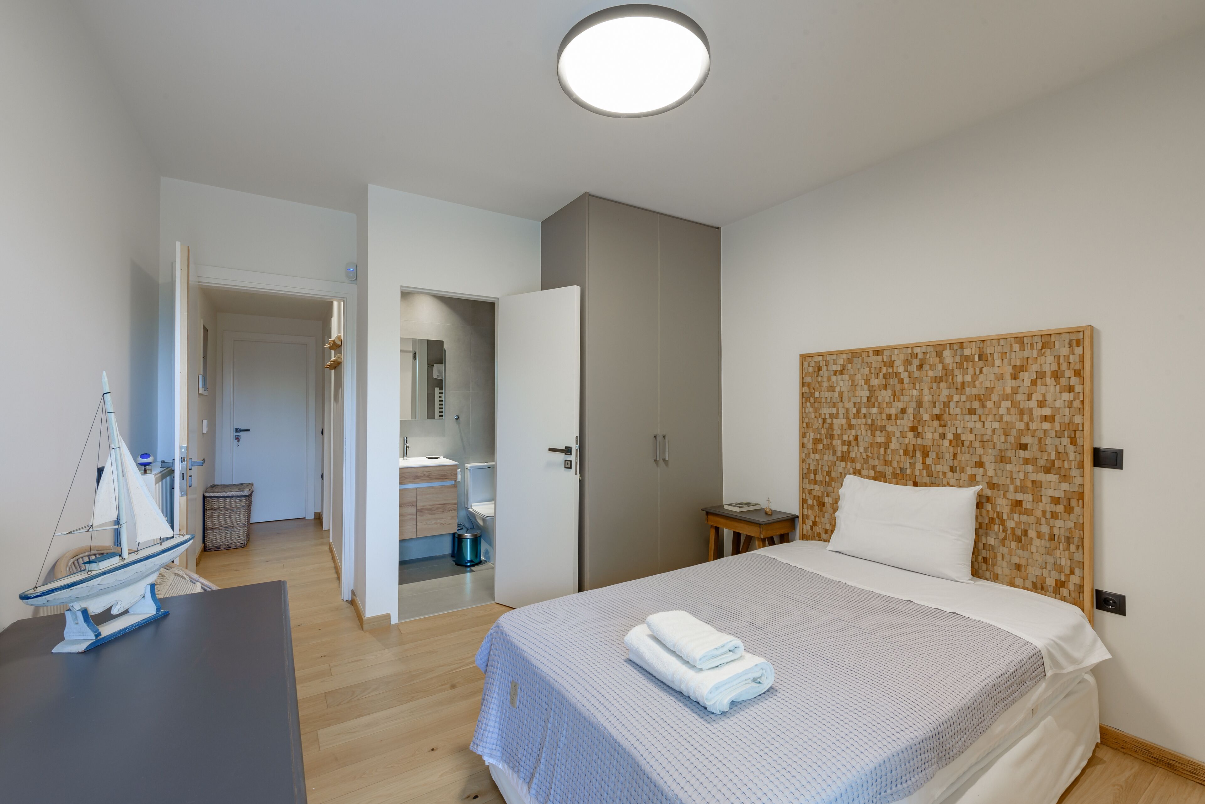 Une chambre d’hôtel avec un lit, une table de chevet, une petite table sur laquelle est posé un modèle de bateau, une porte donnant sur une salle de bain et un luminaire au plafond.