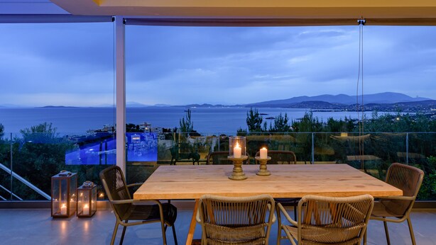 A dining area with a wooden table and wicker chairs, candles, and lanterns, overlooking a scenic view of the sea and hills. - Greek Zen villa - stunning sea view -top of the hill in Athens Riviera (PWV6+C39 Saronida, Greece)