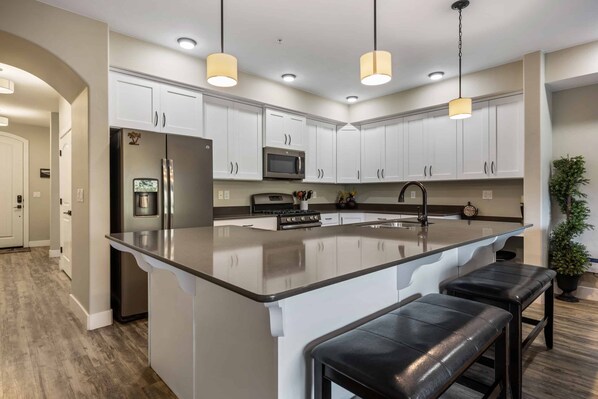 A modern kitchen with white cabinets, a stainless steel refrigerator, a microwave, and a large island with bar stools. - Poolside Palms #W103: Pool, Basketball, Pickleball (St George)