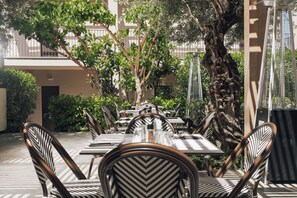 An outdoor dining area with tables and chairs, surrounded by trees and a building facade. - New Beach Hotel (Zakynthos)