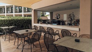 An outdoor seating area with tables and chairs, a bar counter, and a view of a building in the background. - New Beach Hotel (Zakynthos)