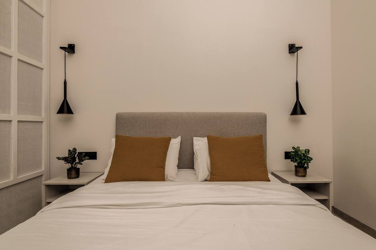 A neatly made bed with white linens, two brown pillows, and a grey headboard. There are two bedside tables with black lamps and potted plants.