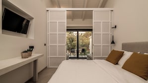 A bedroom with a bed, a TV mounted on the wall, a desk, and a sliding door leading to an outdoor area with chairs. - New Beach Hotel (Zakynthos)