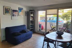 A living room with a blue sofa, a dining table with chairs, a bookshelf, and a sliding glass door leading to a balcony with a view of parked cars and a person outside. - Joli 2 Pièces à L'entrée des Calanques, à Deux Minutes de la Plage (Marseille)