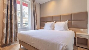 A hotel room with a large bed, a bedside table, a window with curtains, and a view of buildings outside. - Hotel Riviera (Paris)