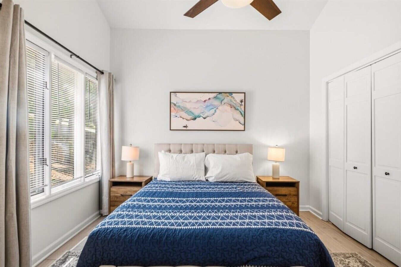 A bedroom with a bed, two bedside lamps, a ceiling fan, a window with blinds, a wall-mounted artwork, and a wardrobe.